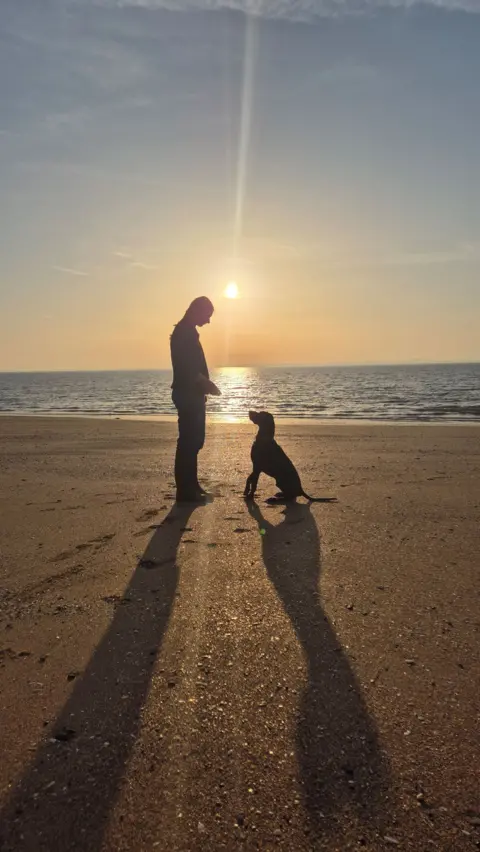 Suzanne Lakie A silhouette of a woman and a dog on a beach. The dog is sitting down looking up at the woman
