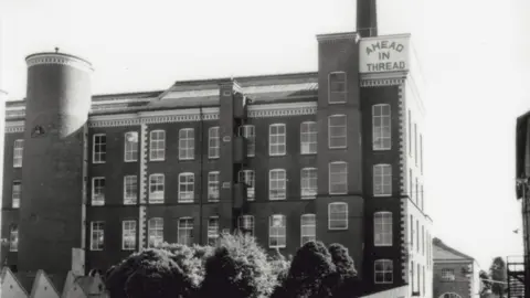 Irish Linen Centre & Lisburn Museum A black and white image of Hilden Mill's spinning mill., which has 'AHEAD IN THREAD' written on a sign at the top. It is a rectangular building with a lot of windows and one circular tower towards the left of the building. The mill can be seen in the background. Trees can be seen along the bottom of the photo.