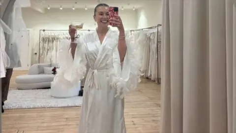 A woman in a large fitting room, wearing a wedding dress and holding a champagne in one hand while taking a selfie photograph