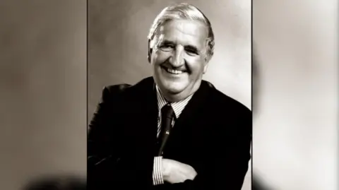 A black and white image of Sir Kenneth Bloomfield smiling at the camera as he wears a suit.