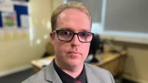 Det Con James Burr sitting in an interview room at Canterbury Police station. He is looking at the camera and wearing dark rimmed glasses, a dark shirt and a grey suit jacket.