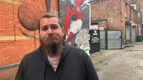 Photograph of Jason Stephenson, who runs the To The Moon & Back bar on Market Avenue in Ashton-under-Lyne. The image shows the Sir Geoff Hurst mural in the background.
