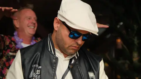 28thStreetMedia A man wearing a white flat cap, sunglasses, a white shirt and a black and white US college-style letterman jacket, DJs. Norman Cook stands in the background watching.