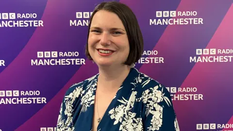 Sarah Wakefield has a blue blouse with white floral pattern. She has dark red bobbed hair.