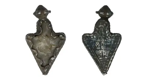 Crown Copyright The amulet is a dark silver colour with a flint inside. The outside of the charm is decorated with carvings.