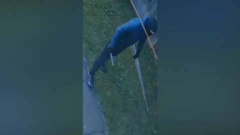 West Yorkshire Police A person wearing a black balaclava, black puffer jacket and jeans stands in a fenced garden. He is holding a silver object in his gloved hand. 