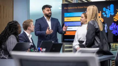 BAE Systems Two men and three women suits having a discussion in what looks like a company boardroom with a large screen behind them.