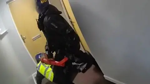 Two armed police - one officer wearing fluorescent jacket and the other in all black - conduct a raid inside a flat corridor. One officer is using a red piece of equipment to knock the door in.