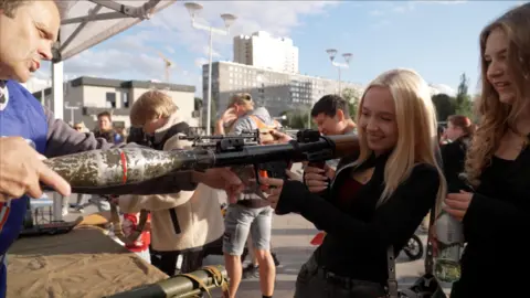 A blonde Russian child in a black top smiles as she holds a rocket launcher, at a weapons demonstration in Moscow
