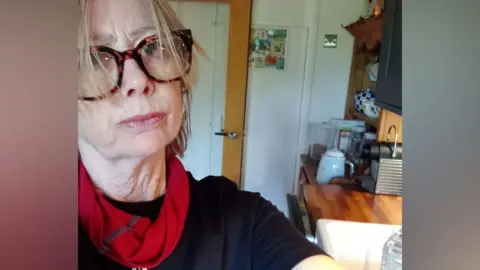 Catherine Catherine stood in a black t-shirt with red scarf and glasses stood in her kitchen.