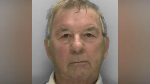 ERSOU A mugshot of an older man. He is looking at the camera, unsmiling. 