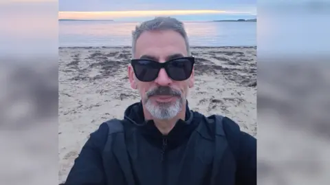 Gavin Hall A man wearing black sunglasses, sporting grey hair and a grey french beard looks into the camera while standing on a beach with the sea behind him.