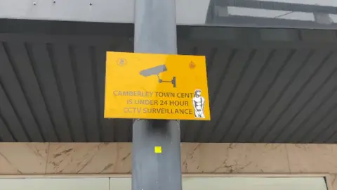 BBC A yellow and black street sign saying Camberley Town Centre is under 24 hour CCTV surveillance.