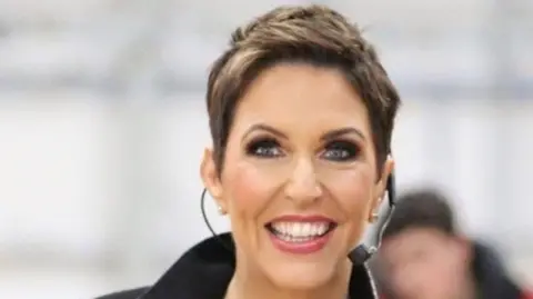 Kendall Glenn Libby Clarke. She has short brown hair and blue eyes, wearing a head microphone.