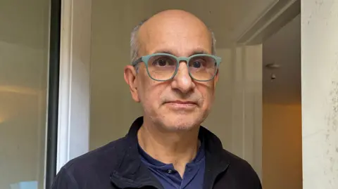 An older man with short grey hair and stubbly grey facial hair stands in his front doorway. He is wearing glasses with a sage green rim, a black fleece and a navy top. The walls inside his flat are white.