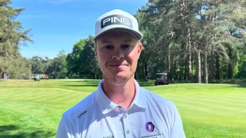 Kate Bradbrook/BBC Brad Smith is smiling at the camera while on the golf course. He is wearing a white cap with the word "Ping" on the front. He is also wearing a white polo shirt with similar branding to what is on his hat. It is a sunny day and the cap is shading his face.