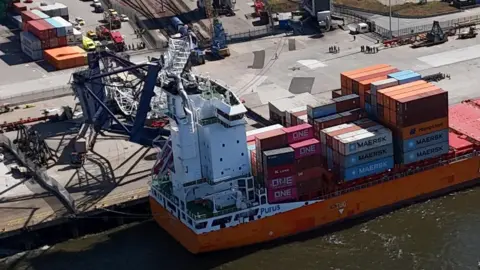 PA Media A crane has been knocked over on its side and is lay on top of an orange container ship. The crane is damaged, as are a number of containers on the vessel. 