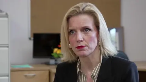 BBC Woman with short blonde hair and red lipstick sat infront of desk in black blazer and striped blouse