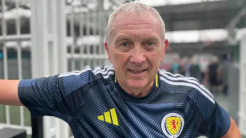 Jim Stewart wearing a blue Scotland top and looking directly at the camera. He is standing in front of a white gate in what appears to be an airport, with the background blurred.