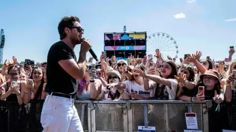 Niall Horan performs at Radio 1's Big Weekend in Sunderland wearing sunglasses, white jeans and black t-shirt. Young female fans behind metal barrier with phones in their hands reach out towards him, arms outstretched. A Ferris wheel is pictured in the background against a blue sky.