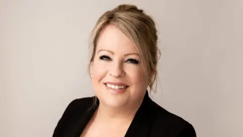 Sharon Livermore Sharon Livermore is standing in front of a beige backdrop. She has blonde hair in a ponytail and is wearing a black blazer and gold earrings.