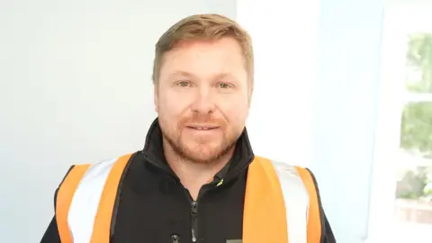 BBC/ Qays Najim A man with light coloured hair and a short moustache and beard stands in a light room. He wears a black jacket and orange high-visibility vest.
