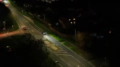 A bird's eye view of the A1079 road going through Hayton at night. Three cars, fairly far apart are seen driving along with their headlights on. Several streetlights are dotted along either side of the road and some houses are visible.