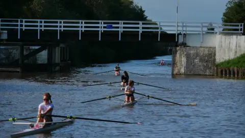 Hartbury College A group of five single skull rowers on a river with a bridge going over them. They are in single file, wearing sorts and t shirts.