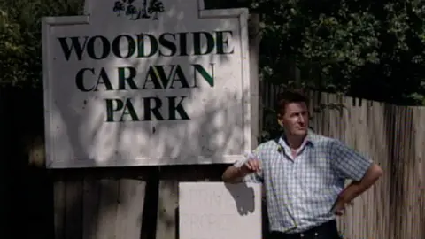 A man in a checked shirt, standing outside on a sunny day. He is standing next to a sign that says "Woodside Caravan Park". One hand is resting on his hip, while the elbow of his other arm is leaning on a smaller sign that faintly says "Private Property". There is a wooden fence and trees behind him.