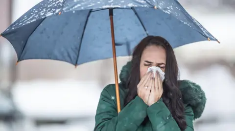 Getty Images Woman sneezing under an umbrella on a cold, wet day