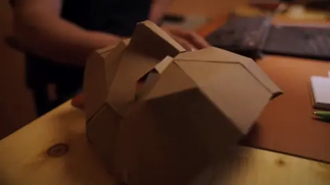 One of the "ninjas" works on his computer, with a paper mask down on his desk.