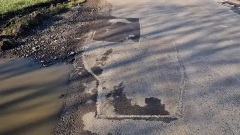 James Coxall A side view of a road showing a large watery puddle to the left and a patched-up pothole in the road to the right