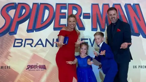 A blond woman in a red dress, man in a black suit, and two children in royal blue do the spider man pose, extending their hands to the camera, in front of the premiere poster.