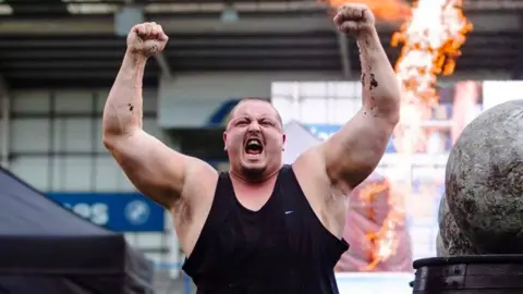 A man with his arms aloft at a strongman competition. He has short hair and is wearing a black vest.