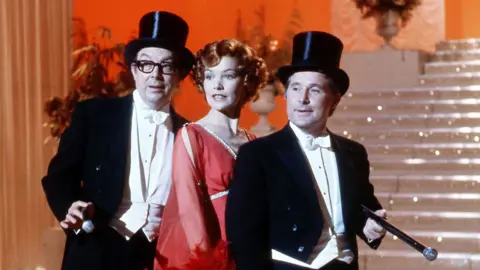 Eric Morecambe, Glenda Jackson, Ernie Wise. The men are in black top hat and tails with white shirts and waistcoats and black sticks tucked under their arms. They flank Jackson who has short curly red hair and a floaty red dress with feathers. The trip stand at the bottom of fairy-lit stairs