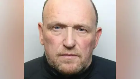 Greater Manchester Police Police custody image of Nicholas Martin. He has receding short grey hair and brown and grey beard and is wearing a black top.