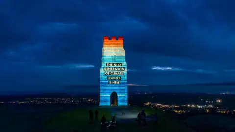 University of Reading Horizontal stripes are projected against the tower on top of Glastonbury Tor. The bottom stripes are mainly dark blue, the middle are mainly light blue and the top are mainly red. It is night time, the sky is filled with moody blue clouds and the town is lit by street lights in the distance.