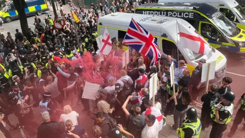 Anti-immigrations protesters waving St George and union jack flags are on one side of a line of police officers on a street. One person is holding a red flare, leaving red smoke above some people's heads. On the other side are counter protesters. There are three police vans to the side of them; the middle one says Heddlu Police and is black.