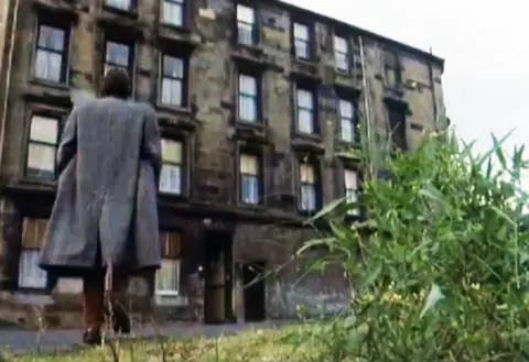 David Hayman, with long hair and a grey overcoat, is walking away from us from some waste ground towards a dirty old tenement building of eight flats.