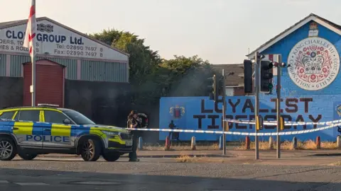 A police officer is behind tape that's cordoning off a road. A police car is parked. A mural saying loyalist Tigersbay is on a wall that's been painted blue.
