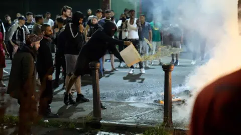 Dan Nelson/BBC A man in a black hooded top and face mask throws a bucket of water towards something on fire, which is out of the picture. He is surrounded by a crowd of people. 