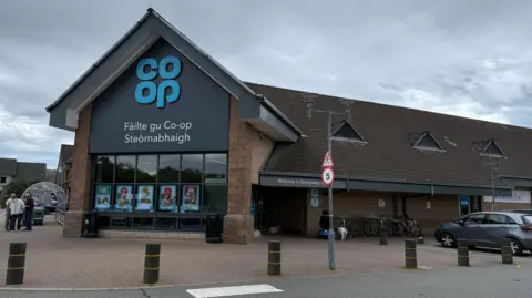 BBC The Co-op in Stornoway is a large building. There is a front entrance with a glass frontage and peaked roof. The word "Co-op" is in blue letters and below it words in Gaelic to say it is the Co-op in Stornoway. Behind the entrance is a long building with a peaked roof.
