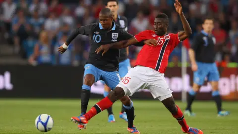Getty Images Two male football players duel for the ball in open play, with an England player in black and blue kit losing possession to a Norway player in red and white kit, with other players, the pitch and spectators seen in soft focus in the background.