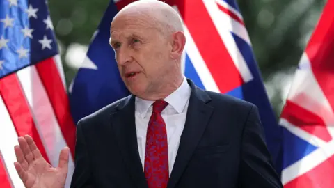 John Healey standing in front of the flags of the US, UK and Australia 