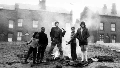 A black and white photo of a group of boys and one girl stood next to a small fire on a mound of grass. Some terraced houses can be seen in the background.