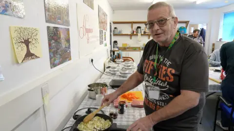 Martyn stands in front of a hob, stirring onions in a frying pan. He has short greying hair. He's wearing a glasses, a green sunflower lanyard, and a grey t-shirt bearing the slogan "born in the fifties" in white and orange lettering. In the background, pie ingredients (mince blocks and chopped carrots) are visible, as well as 6 pie-making machines. 