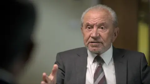 Alan Sugar wears a grey suit jacket, white shirt and striped tie; he has short grey hair