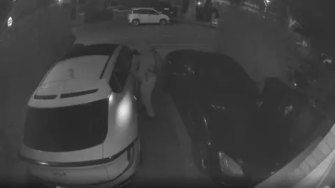 A hooded man appears to be tampering with a white Kia car parked outside a home, the picture is taken on a doorbell camera