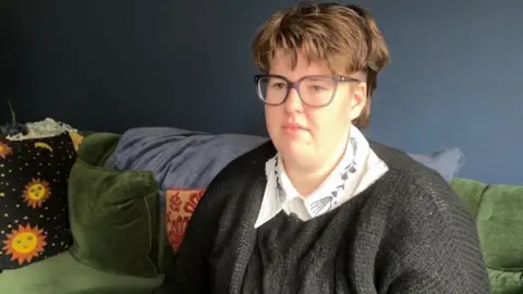 Leah Duncan is sitting on her green couch ad looking at the interviewer behind the camera. She has short brown hair and glasses.