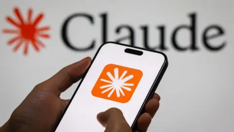 A close up of a hand holding a smartphone displaying Anthropic's Claude logo on a white screen. The logo is displayed in the background of the image behind it.